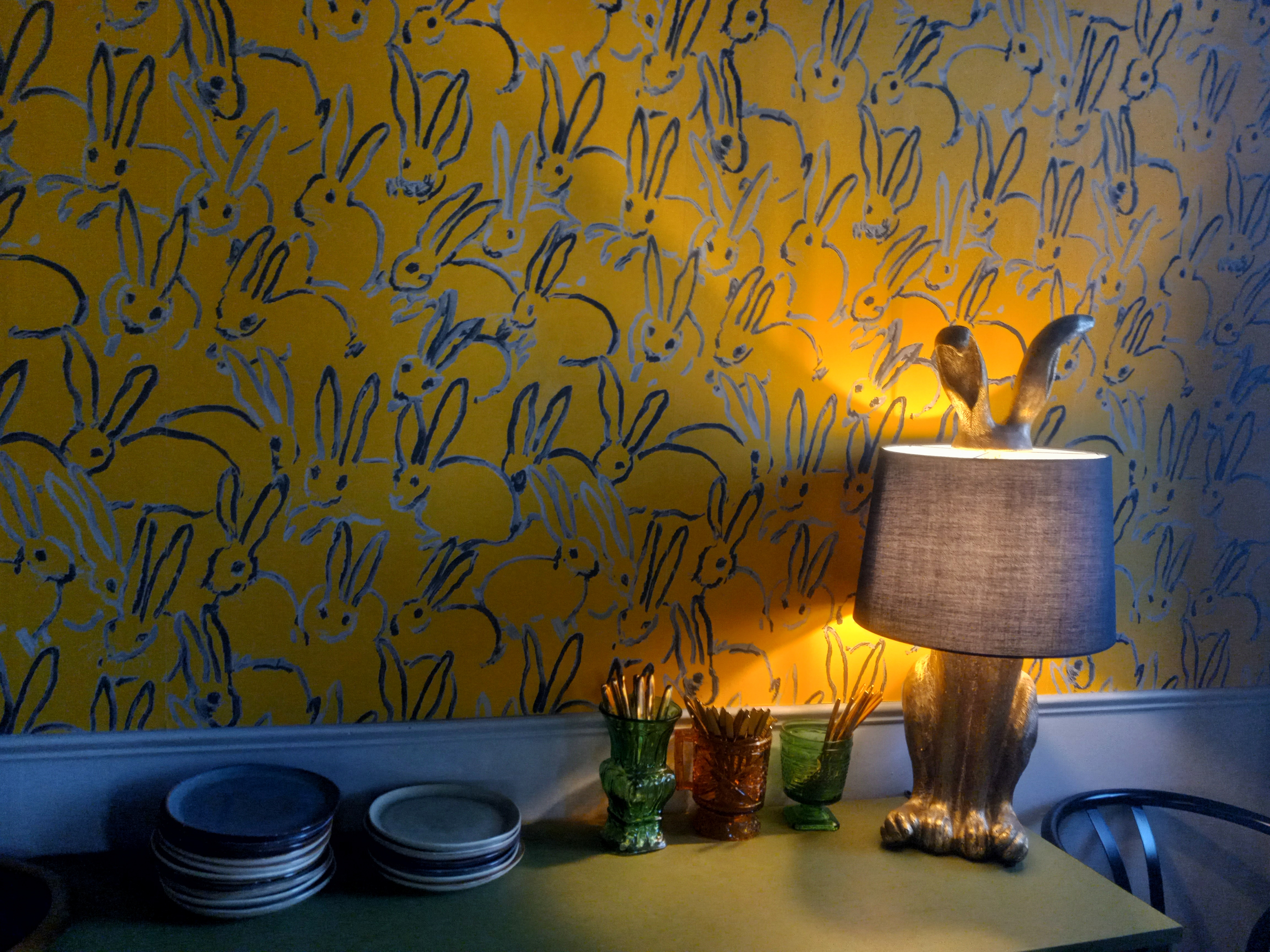 A dark yellow wall covered with paintings of blue rabbits. In front of the wall is a table with a rabbit-shaped lamp on it