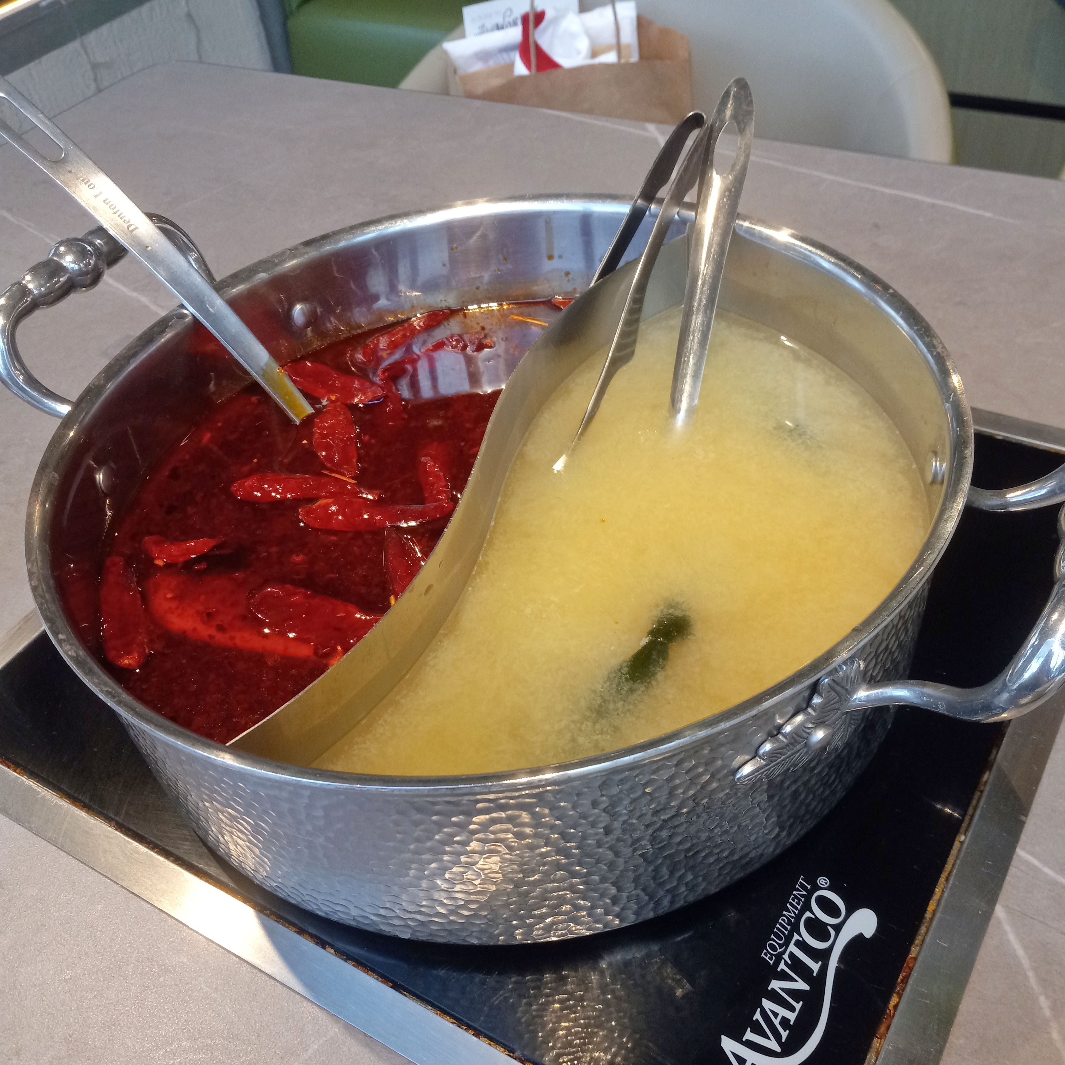 A divided pot filled with two kinds of broth. One broth is pale yellow and the other is bright red with chili peppers